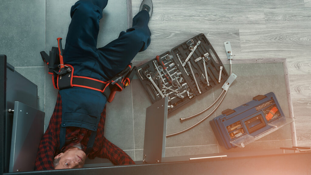 High angle view of plumber in dunwoody wearing auniform lying on floor in the kitchen and using wrench while repairing water pipe in kitchen. Toolbox on the floor. Horizontal shot
